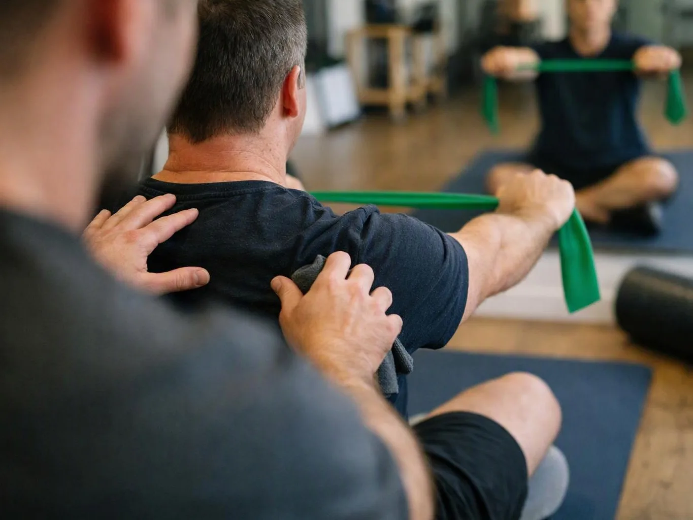 A therapist helps a seated man stretch with a green resistance band in a gym studio